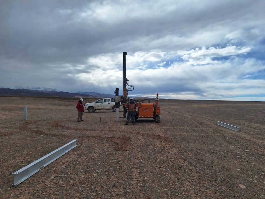 2 operadores junto a máquina hinicadora de perfiles metálicos delante de camioneta blanca en obra en desierto